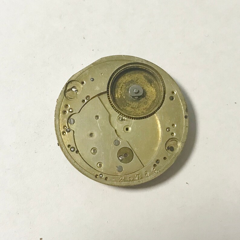 May include: Close-up of a gold-toned watch movement with intricate gears and a circular winding mechanism. The movement is set against a white background.