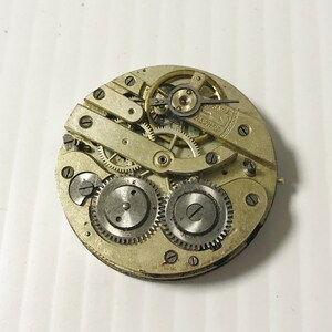 Puede incluir: Primer plano de un movimiento de reloj de bolsillo plateado con engranajes intrincados y un v&aacute;stago de cuerda. La esfera del reloj no es visible.