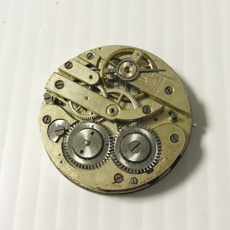 Puede incluir: Primer plano de un movimiento de reloj de bolsillo plateado con engranajes intrincados y un v&aacute;stago de cuerda. La esfera del reloj no es visible.
