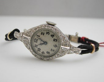 May include: A vintage wristwatch with a silver face and diamond accents. The watch has a black leather band and a white dial with black Roman numeral hour markers.