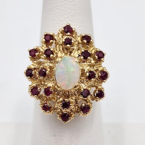 May include: A gold-toned ring featuring an oval opal center stone surrounded by a cluster of small, round, deep red gemstones. The ring's setting has a textured, floral-like design.