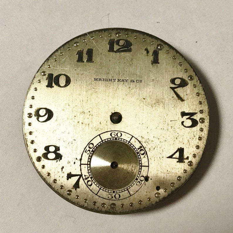 May include: A silver watch face with black Roman numeral hour markers and black Arabic numeral minute markers. The watch face has a small, circular subdial with black Arabic numeral markings. The text "WRIGHT KAY & CO" is printed on the watch face.