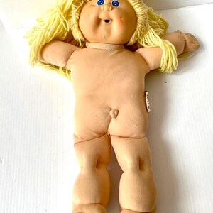 May include: A vintage Cabbage Patch Kid doll with blonde yarn hair and blue eyes. The doll is wearing a white shirt and has a pink blush on its cheeks.