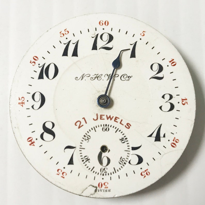 May include: A white pocket watch face with black Roman numeral hour markers and black Arabic numeral minute markers. The watch face has a black hand and a silver second hand. The watch face also has the text "N.F.C. W. Co." and "21 Jewels" in black text. The watch face is surrounded by a white metal case with a silver rim.