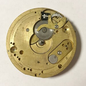 May include: Close-up of a gold-toned watch movement with intricate gears and a silver-toned lever. The movement is mounted on a brass plate with a circular shape.
