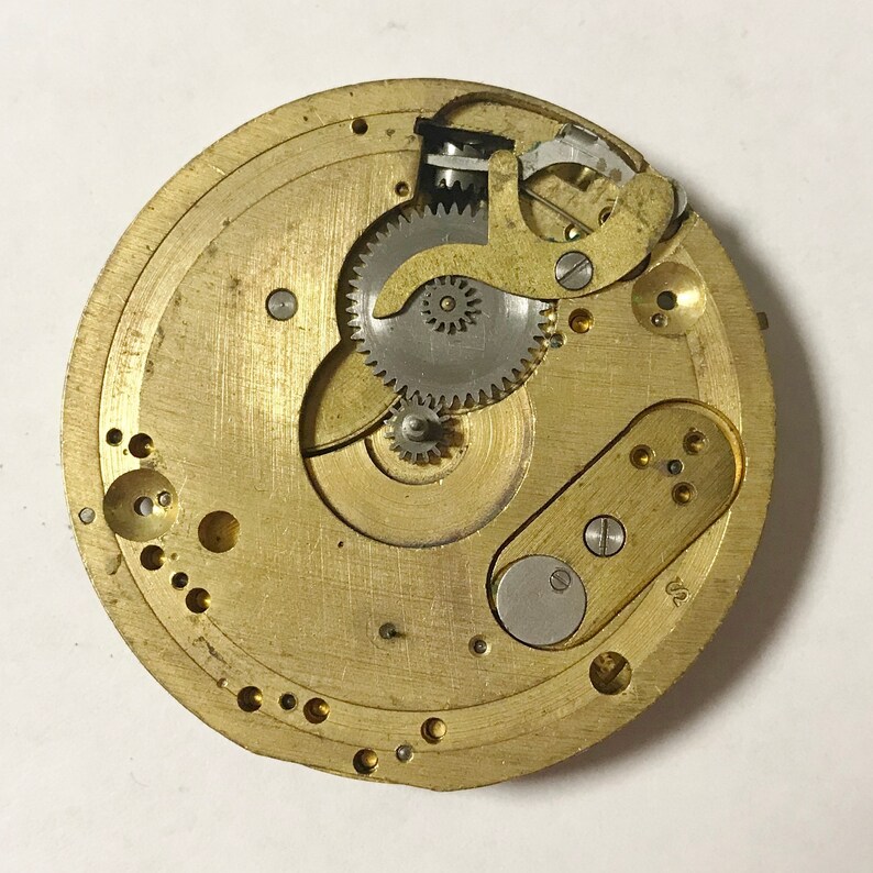 May include: Close-up of a gold-toned watch movement with intricate gears and a silver-toned lever. The movement is mounted on a brass plate with a circular shape.