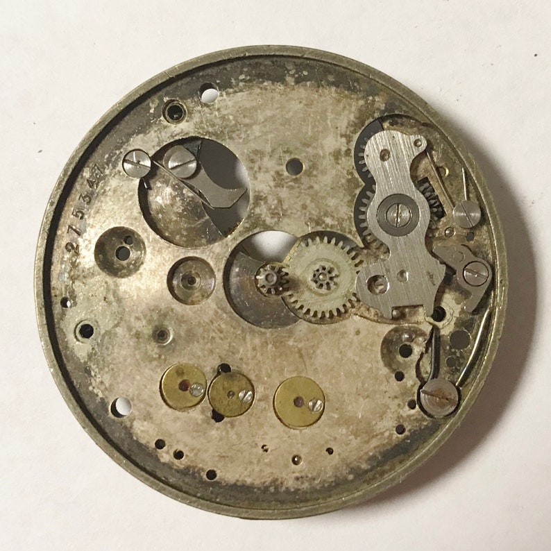 Pu&ograve; includere: Vista ravvicinata del meccanismo interno di un orologio argentato. Gli ingranaggi e le leve intricate sono visibili, insieme al numero "27534" stampato sul lato.