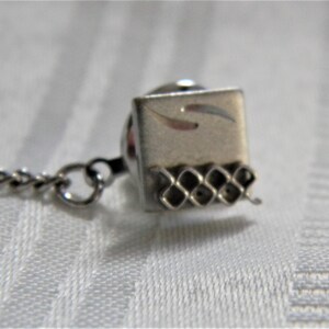May include: Silver tie tack with a square design and a chain. The square has a decorative etched pattern and a small chain attached.