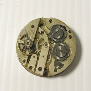 Puede incluir: Primer plano de un movimiento de reloj plateado con varios engranajes y un v&aacute;stago de cuerda. El movimiento del reloj est&aacute; expuesto y muestra el funcionamiento intrincado del reloj.