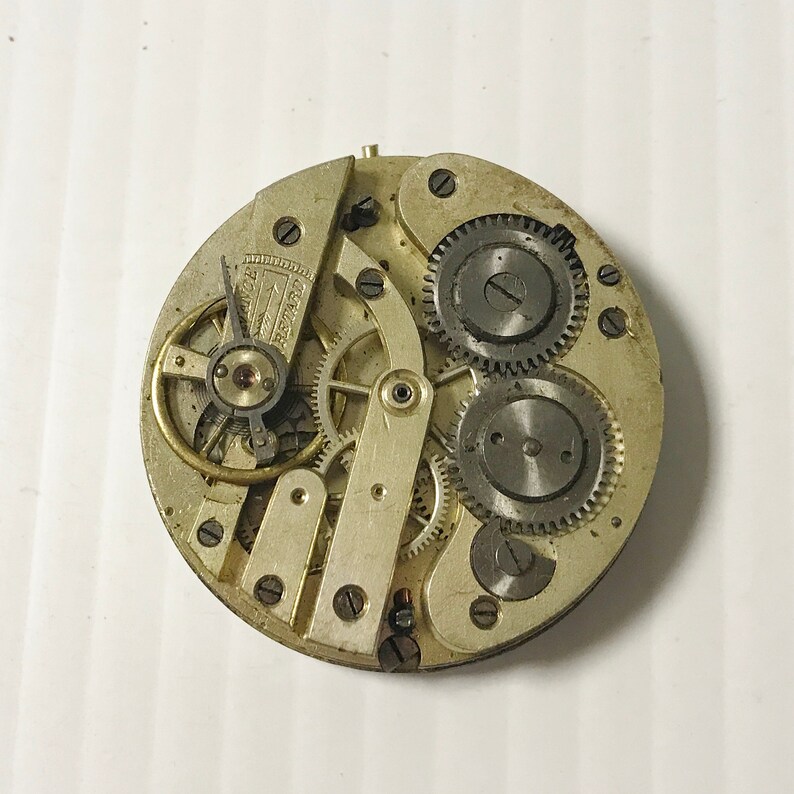 Puede incluir: Primer plano de un movimiento de reloj plateado con varios engranajes y un v&aacute;stago de cuerda. El movimiento del reloj est&aacute; expuesto y muestra el funcionamiento intrincado del reloj.