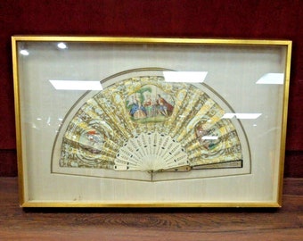 May include: A framed antique fan with a gold and white design. The fan is decorated with a painted scene of people in a garden.