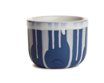 May include: A ceramic cup with a rounded base and a white interior. The exterior is navy blue with white vertical drip patterns. The rim is white with a blue accent. The bottom of the cup has the word "KAYE".