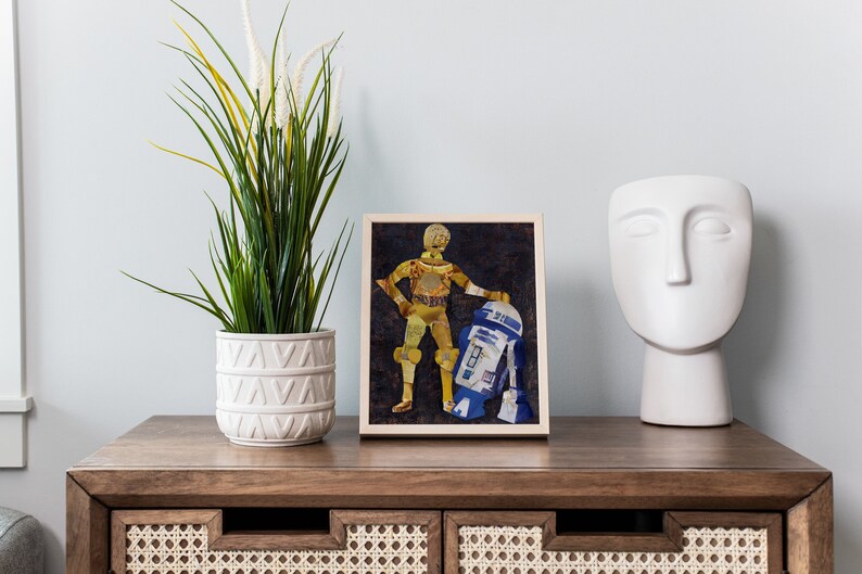 May include: A framed print of a Star Wars character, C-3PO, standing next to R2-D2. The print is made of paper and has a collage-style design. The print is on a wooden table with a white vase and a white sculpture.