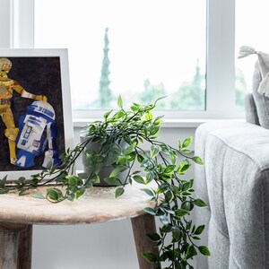 May include: A framed collage art print of C-3PO and R2-D2 from Star Wars. The characters are made of recycled materials and are displayed on a dark brown background. The frame is white and the print is sitting on a wooden table.