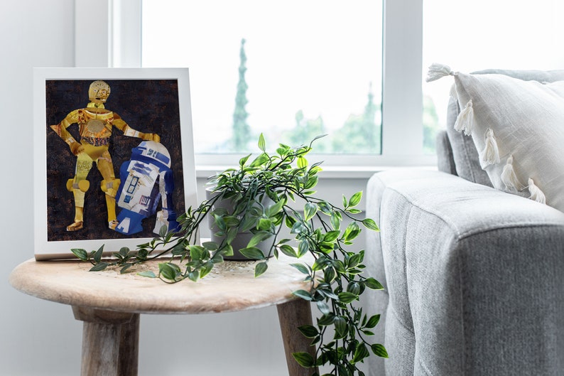 May include: A framed collage art print of C-3PO and R2-D2 from Star Wars. The characters are made of recycled materials and are displayed on a dark brown background. The frame is white and the print is sitting on a wooden table.