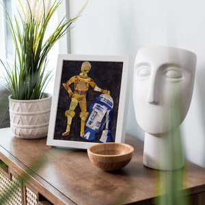 May include: A framed collage of Star Wars characters C-3PO and R2-D2 made from recycled materials, displayed on a wooden table with a white ceramic sculpture and a potted plant.