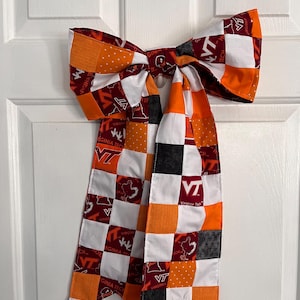 May include: A large, handmade bow with a patchwork design in maroon, orange, white, and black. The bow features the Virginia Tech logo and text. The bow is hanging on a white door.