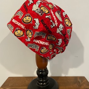 Peut inclure: Un bonnet de chirurgien en tissu rouge avec un motif répété du logo et de la mascotte des Ohio State Buckeyes. Le bonnet est sur un support en bois.