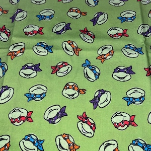 May include: Green fabric featuring a repeating pattern of cartoon turtle faces with different colored masks: red, orange, blue, and purple. The turtles have white shells and are smiling. The background is a light green color.