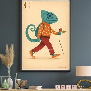May include: A framed art print featuring a whimsical illustration of a teal chameleon wearing an orange sweater with white polka dots, red pants, and orange shoes. The chameleon carries a ladybug on a stem. The letter "C" is in the upper left corner.