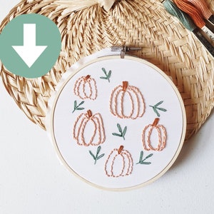 May include: Embroidery hoop featuring a pumpkin design with orange thread and green leaves. The hoop is set against a woven, heart-shaped background with spools of thread in matching colors. A craft project with a fall theme.