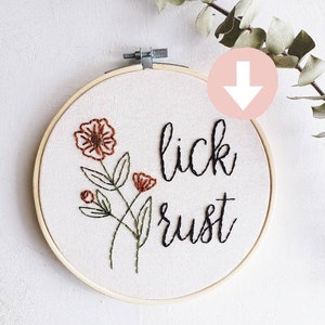 May include: Embroidered hoop art with the words "lick rust" in black script. The design includes embroidered flowers in brown and green. The artwork is set in a wooden embroidery hoop.