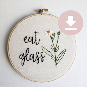 May include: Embroidered artwork in a wooden hoop. The words "eat glass" are stitched in black on off-white fabric, with a daisy and green leaves. The hoop is secured with a brass screw.