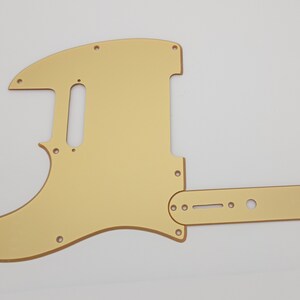 May include: Gold-colored guitar pickguard with a rectangular control plate. The pickguard has a rounded shape and several holes for screws and knobs.