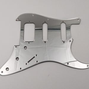 May include: A polished, chrome guitar pickguard with various screw holes and apertures for pickups and controls. The pickguard has a contoured shape, designed for a particular guitar. The surface reflects light, emphasising its smooth finish and metallic appearance.
