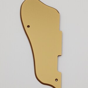 May include: Gold-colored guitar pickguard with a rounded shape and two small holes.