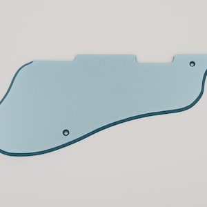 May include: A light blue guitar pickguard with a curved, asymmetrical shape. The pickguard has two small holes for mounting and a dark blue outline. The object is set against a white background.