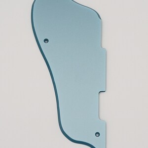 May include: A light blue guitar pickguard with a unique shape, featuring two screw holes and a cutout. The pickguard has a smooth surface and is designed to protect the guitar's body from scratches. It is a replacement part for an electric guitar.