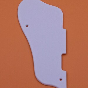 May include: A white plastic guitar pickguard with two small holes. The pickguard is shaped like a traditional guitar pickguard and is designed to fit a standard electric guitar.
