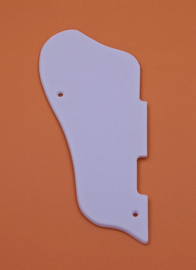 May include: A white plastic guitar pickguard with two small holes. The pickguard is shaped like a traditional guitar pickguard and is designed to fit a standard electric guitar.