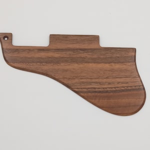 Può includere: Un battipenna per chitarra in legno marrone, dalla forma unica, con un bordo inferiore curvo e un piccolo foro nella parte superiore. La venatura del legno è visibile, aggiungendo consistenza a questo accessorio per strumenti.
