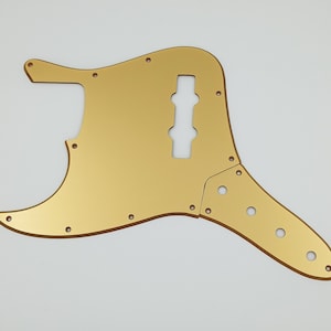 Könnte beinhalten: Ein goldfarbenes Gitarren-Schlagbrett mit mehreren Schraubenlöchern und einem Ausschnitt für Tonabnehmer. Das Schlagbrett hat eine glatte, reflektierende Oberfläche und ist für ein bestimmtes Gitarrenmodell konzipiert. Die Gesamtform ist asymmetrisch.