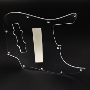 Könnte beinhalten: Transparente Plastik-Pickguard für einen Bass. Das Pickguard hat einen Ausschnitt für die Reglerknöpfe und eine rechteckige Öffnung für einen Tonabnehmer.