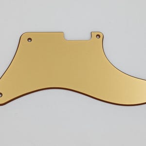 Op de afbeelding: Een goudkleurige gitaar pickguard met een unieke vorm, met drie schroefgaten aan de onderkant en twee aan de bovenkant. De pickguard heeft een glad, reflecterend oppervlak en is ontworpen om de gitaarbody te beschermen.