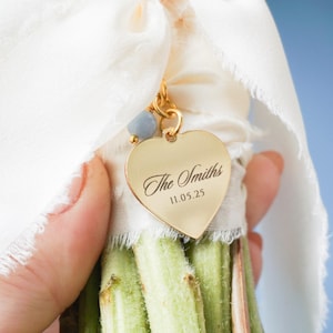 May include: A bouquet of green stems tied with a white ribbon and a gold heart-shaped charm. The charm is engraved with "The Smiths" and "11.05.25". A small blue bead hangs from the charm. The background is a soft blue.