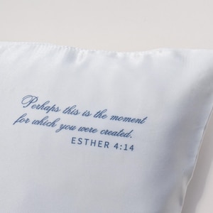 May include: A light blue pillow with the quote "Perhaps this is the moment for which you were created. ESTHER 4:14" printed in a dark blue script. The pillow has a smooth, satin-like texture.