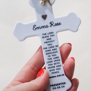 May include: A white ceramic cross ornament featuring the name Emma Rose, a small heart, and the text of Numbers 6:24-26, finished with a date of 06.08.26. It includes a dusty rose velvet ribbon and a gold-toned cross and pearl charm attachment.