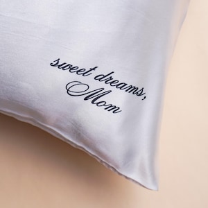 May include: A light gray satin pillow with the words "sweet dreams, Mom" in a cursive font. The pillow has a soft, smooth texture and a subtle sheen, suggesting a luxurious feel. The text is in a dark blue color.