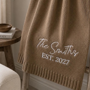 May include: A solid brown knitted throw blanket featuring white embroidered text that reads The Smiths and EST. 2027. The blanket has a ribbed border at the bottom edge and is draped over a wooden ladder, showcasing its textured knit construction.