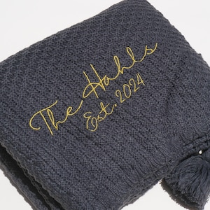 May include: A folded, dark gray knitted blanket with a textured pattern. The blanket has a yellow embroidered script that reads "The Hahls Est. 2024". A tassel hangs from the corner.