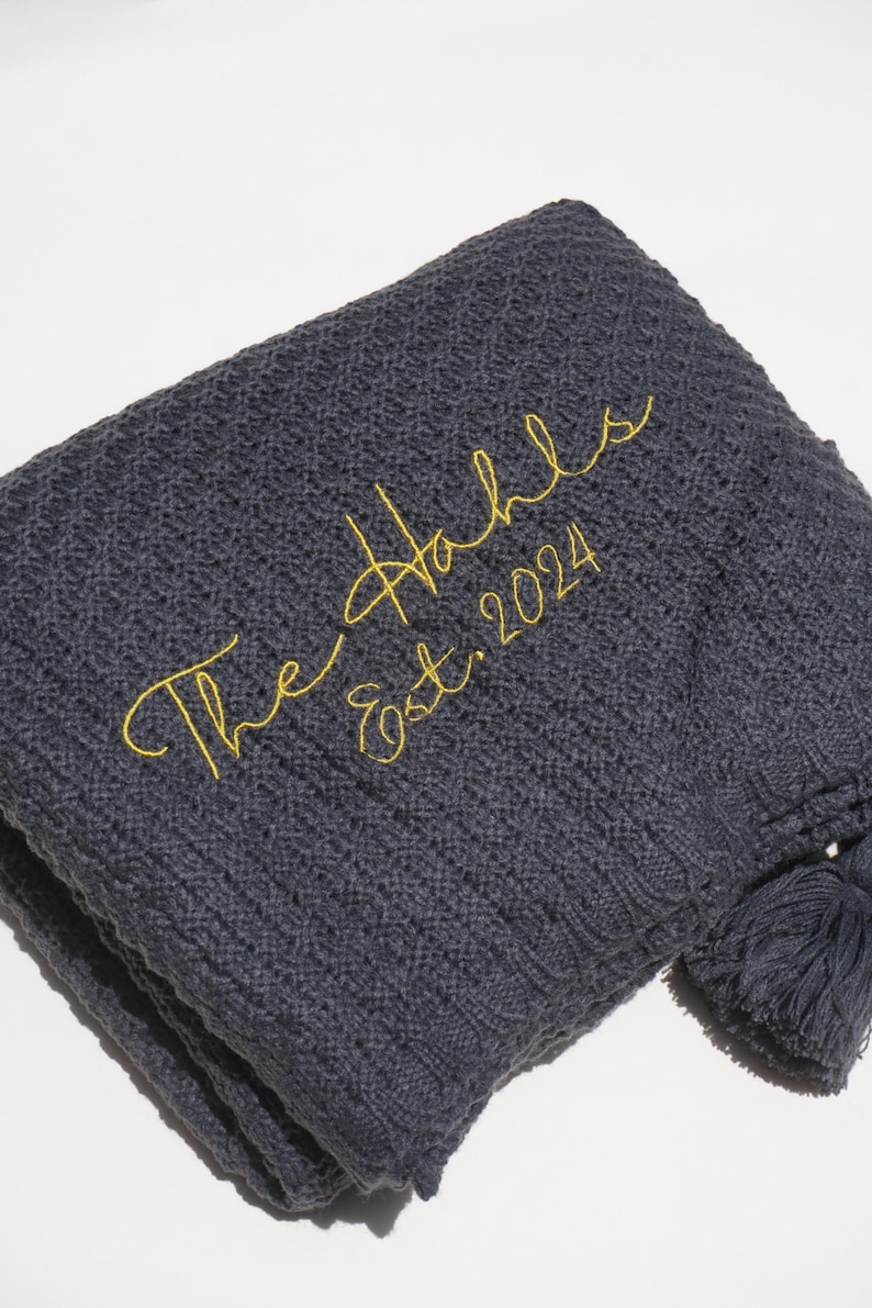 May include: A folded, dark gray knitted blanket with a textured pattern. The blanket has a yellow embroidered script that reads "The Hahls Est. 2024". A tassel hangs from the corner.