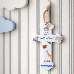 May include: A white ceramic cross ornament featuring a colorful rainbow, Noah's ark, and various animal illustrations. The text reads GOD BLESS, Nathan Paul, and BAPTIZED 05.10.26. It is suspended by a light beige fabric ribbon for hanging display.