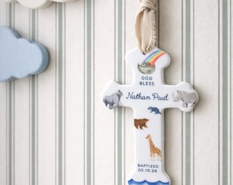May include: A white ceramic cross ornament featuring a colorful rainbow, Noah's ark, and various animal illustrations. The text reads GOD BLESS, Nathan Paul, and BAPTIZED 05.10.26. It is suspended by a light beige fabric ribbon for hanging display.