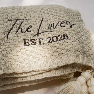 May include: Cream-colored knitted blanket with the embroidered text "The Loves EST. 2026". The blanket has a textured pattern and is folded, with tassels visible on the edge. A cozy home decor item.