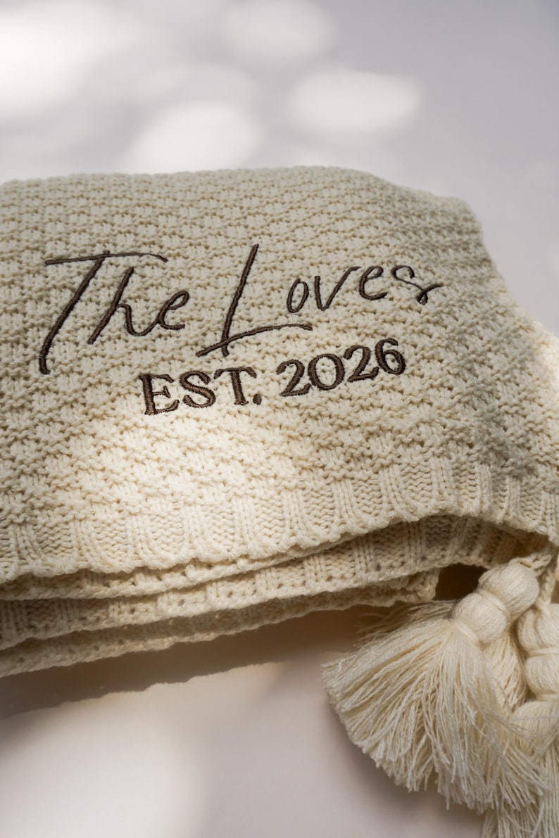 May include: Cream-colored knitted blanket with the embroidered text "The Loves EST. 2026". The blanket has a textured pattern and is folded, with tassels visible on the edge. A cozy home decor item.