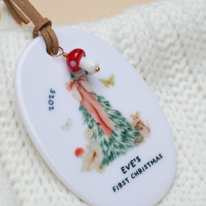 May include: White oval Christmas ornament with a painted Christmas tree design, butterflies, and the text "EVE'S FIRST CHRISTMAS 2025." A red and white mushroom bead hangs from a brown leather strap.